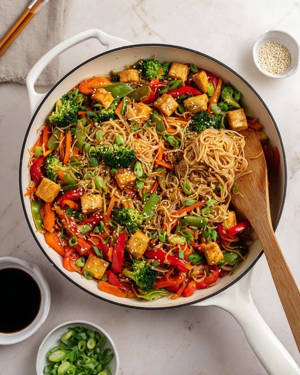 The image shows a white-handled black skillet filled with stir-fried noodles mixed with colorful vegetables and tofu. The bottom layer consists of light brown noodles swirling throughout the pan. On top of the noodles are bright green broccoli florets and snap peas, thin orange carrot slices, and red bell pepper strips. Small golden-brown tofu cubes are scattered generously over the vegetables. The dish is garnished with white sesame seeds and fresh green chopped herbs. A wooden spatula rests inside the pan on the right side. The skillet sits on a white marbled surface, with a white plate and wooden chopsticks at the top left corner, a small white bowl of chopped green herbs in the top right, and a scalloped white bowl of dark sauce and a bowl of sesame seeds near the bottom right. photo taken with an iphone --ar 4:5 --v 7