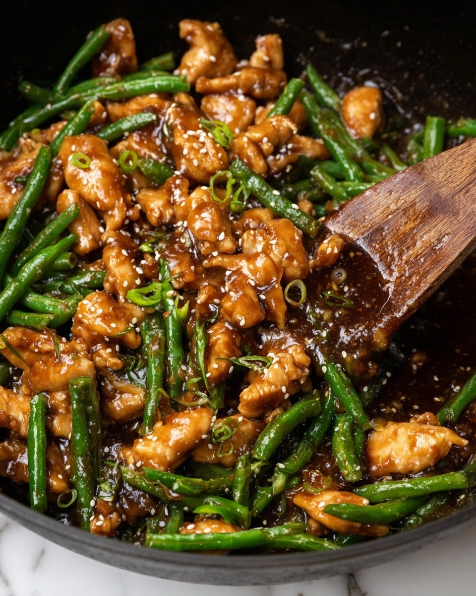 The image shows a close-up of a stir-fry dish in a black pan with pieces of cooked chicken in a shiny brown sauce mixed with bright green cut green beans. The chicken pieces are irregular and coated well in the sauce, which is thick and glossy. Small white sesame seeds are sprinkled throughout, adding texture and contrast to the green beans and chicken. There are also small green onion pieces scattered on top, adding fresh green spots. A wooden spoon is lifting part of the mixture, showing the glossy sauce dripping and clinging to the ingredients. The background is a white marbled texture. Photo taken with an iphone --ar 4:5 --v 7