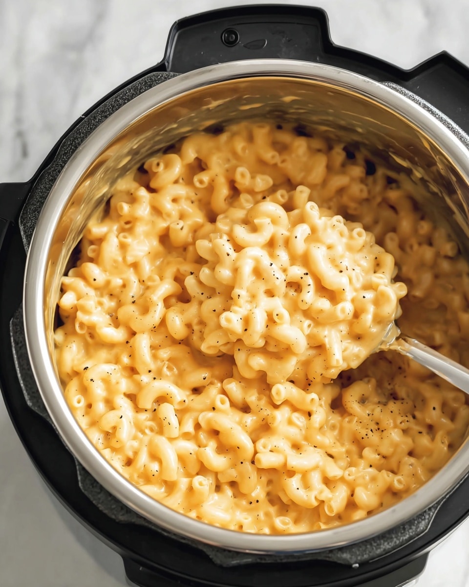 The image shows a close-up of creamy macaroni and cheese inside a shiny silver stainless steel pot with a black handle and base, likely an electric cooker. The dish has one main layer of soft elbow macaroni pasta coated evenly in a smooth, orange-yellow cheese sauce with small black pepper specks scattered on top. A silver spoon is partly inside the pot, lifting a scoop of the cheesy pasta. The background is a white marbled surface. photo taken with an iphone --ar 4:5 --v 7