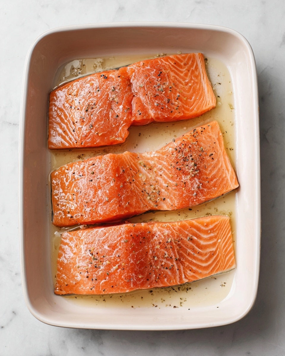 Four pieces of raw salmon fillets with a shiny, wet texture lie flat in a white ceramic baking dish. Each piece is orange with natural salmon lines and is lightly sprinkled with black pepper and small bits of seasoning. The fillets are arranged in two rows with two pieces in each row, fitting neatly within the dish. A small amount of oil gives a slight gloss on the salmon’s surface and the dish bottom. The dish rests on a white marbled surface. Photo taken with an iphone --ar 4:5 --v 7