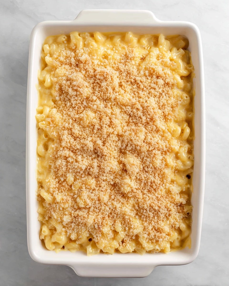 A white rectangular baking dish filled with a creamy macaroni and cheese casserole. The dish has a thick layer of elbow macaroni covered in smooth, melted yellow cheese sauce. On top, a light golden brown crumb layer is spread evenly, adding texture to the surface. The dish is set on a white marbled texture background. photo taken with an iphone --ar 4:5 --v 7