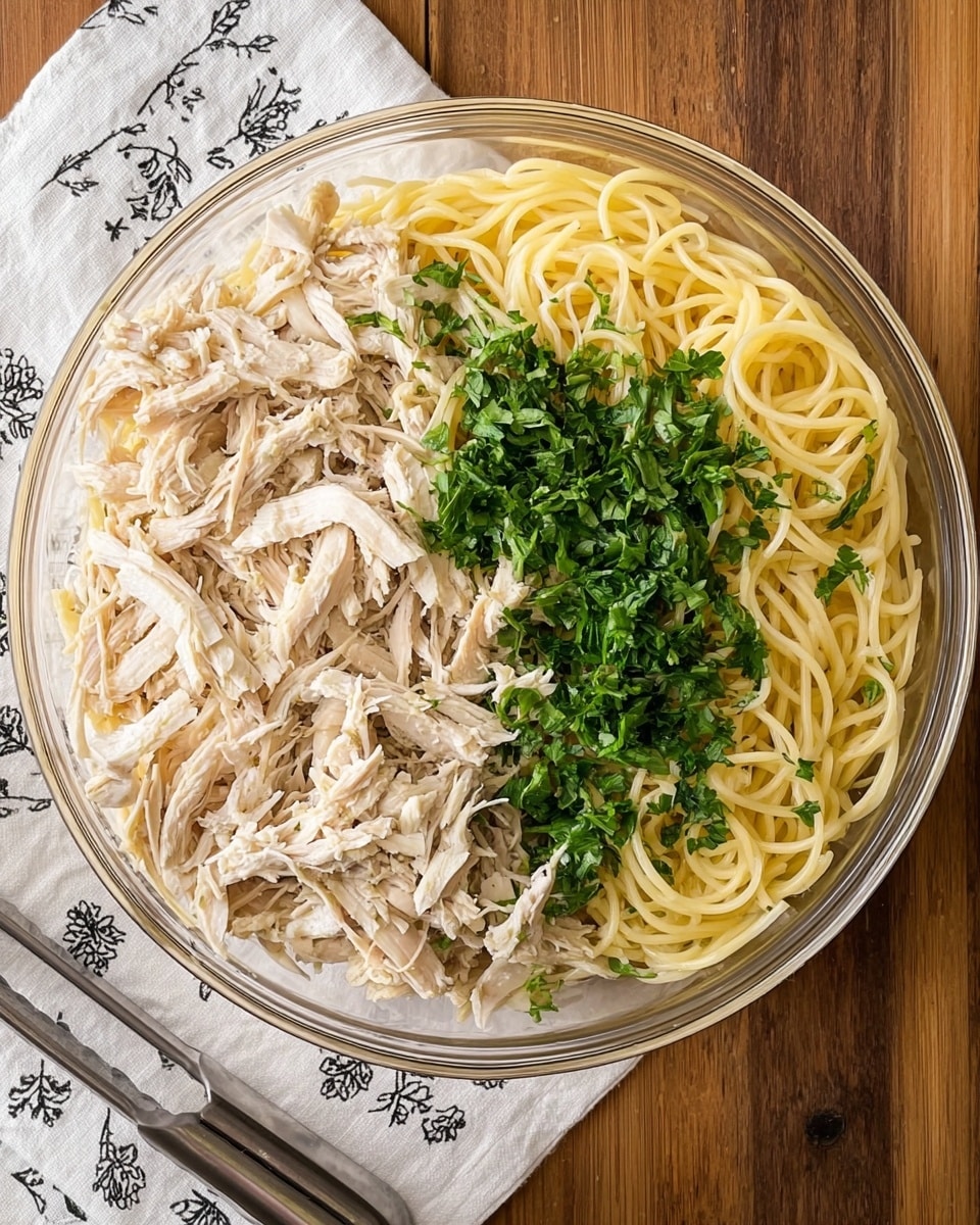 A clear round bowl shows two main halves of food inside: on the left side, shredded light beige chicken with a soft and fibrous texture, and on the right side, cooked pale yellow spaghetti noodles that are smooth and slightly shiny. In the middle, fresh bright green chopped parsley is spread over the border where chicken and noodles meet. The bowl sits on a wooden surface, partly covered by a white cloth with a small black floral print. A pair of metal tongs with black handles rests at the bottom left corner. Photo taken with an iphone --ar 4:5 --v 7