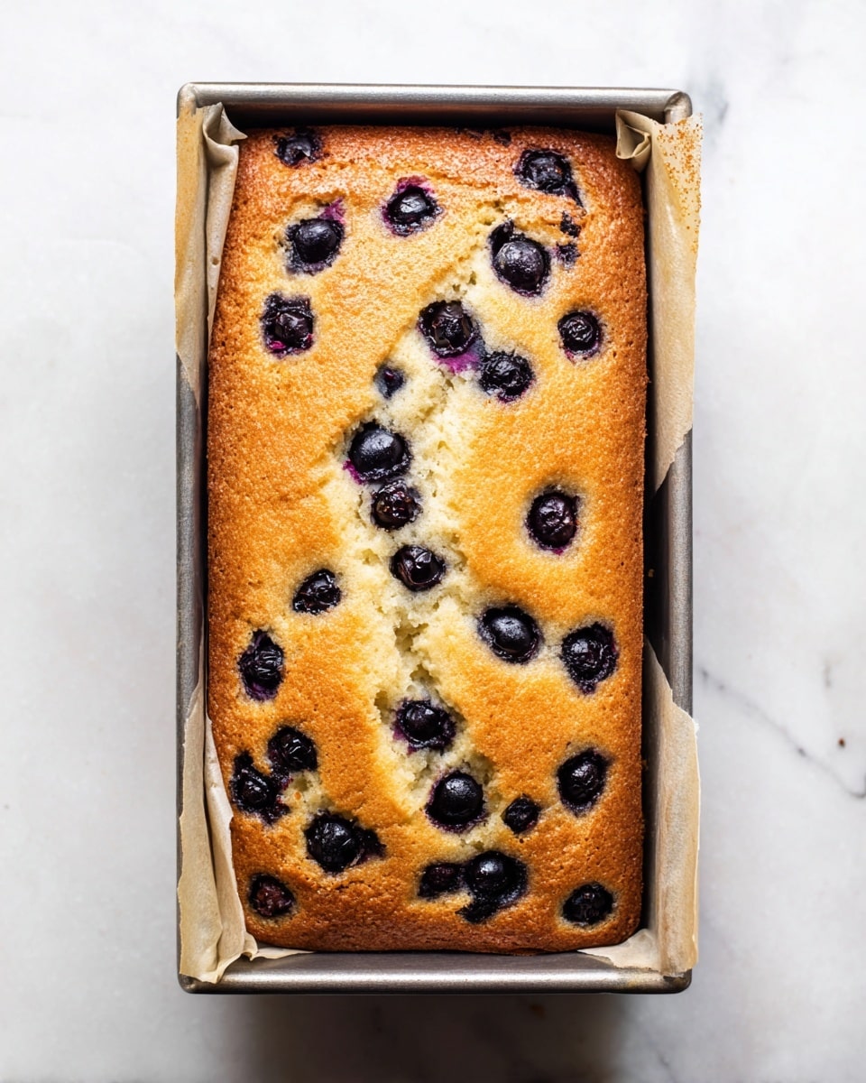 A rectangular baked blueberry cake with a golden brown top layer, showing baked blueberries scattered unevenly across the surface. The cake appears soft and moist with cracks running through the top, and it is still in a silver loaf pan lined with parchment paper on a white marbled surface. The edges of the cake are slightly darker and well-baked, creating a contrast with the lighter middle area photo taken with an iphone --ar 4:5 --v 7