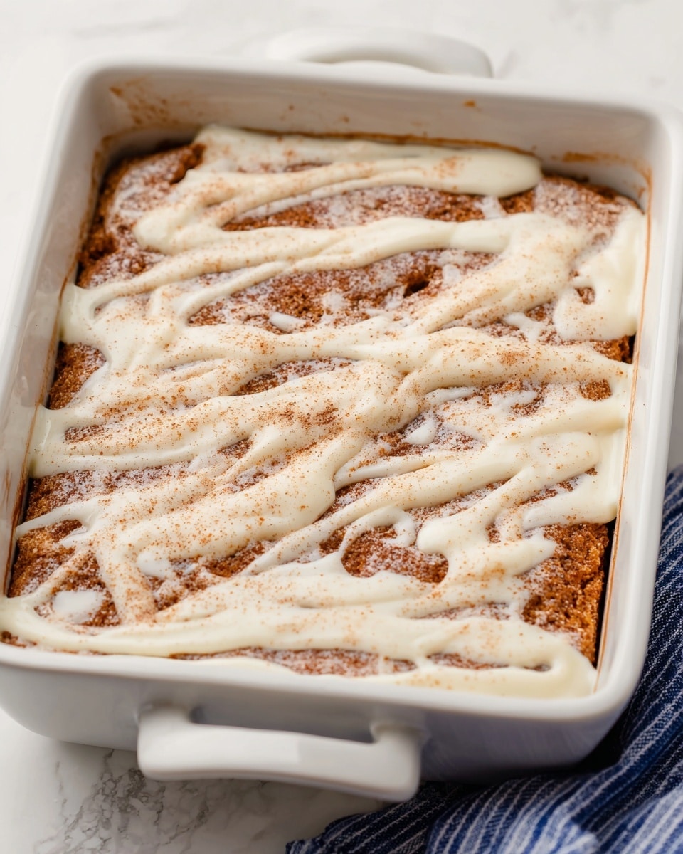 A close-up of a baked dessert in a white rectangular baking dish with two handles visible, featuring one thick baked layer with a golden brown surface sprinkled with cinnamon or sugar, topped unevenly with a creamy white icing drizzled in a flowing pattern across the entire surface. The dish sits on a white marbled texture surface, and a blue and white striped fabric is partially visible at the bottom right corner. photo taken with an iphone --ar 4:5 --v 7