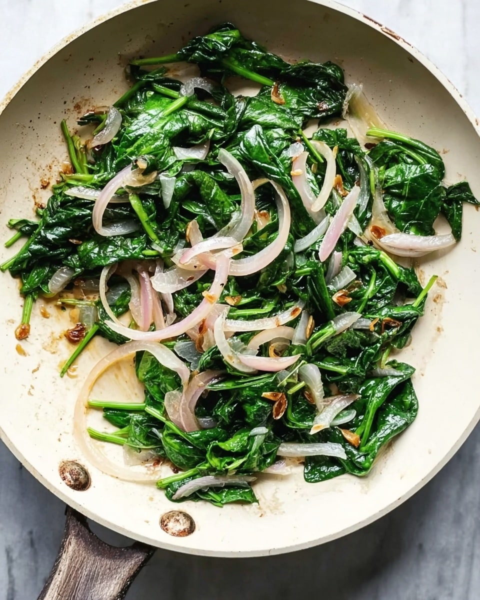 A white frying pan on a white marbled surface holds cooked spinach and sliced onions. The spinach is a deep green color, wilted but still fresh, mixed with light pinkish-white onion strips, some showing light browning from cooking. Small pieces of brown toasted garlic are scattered throughout. The pan's surface shows slight oil gloss. photo taken with an iphone --ar 4:5 --v 7
