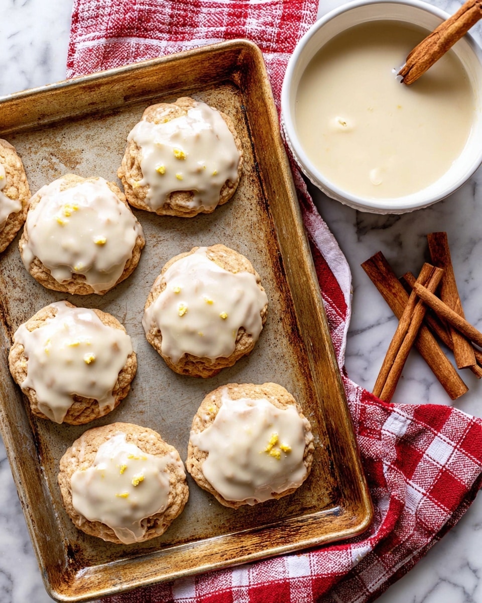 The image shows a metal baking tray with eight round cookies arranged loosely, each topped with a thick layer of pale cream-colored glaze that drips slightly over the edges. The cookies themselves are light brown with a slightly rough texture and small yellow bits visible under the glaze. To the side, there is a white bowl filled with more glaze on a red and white checkered cloth, along with three cinnamon sticks placed on the same cloth, all set on a white marbled surface. Photo taken with an iphone --ar 4:5 --v 7