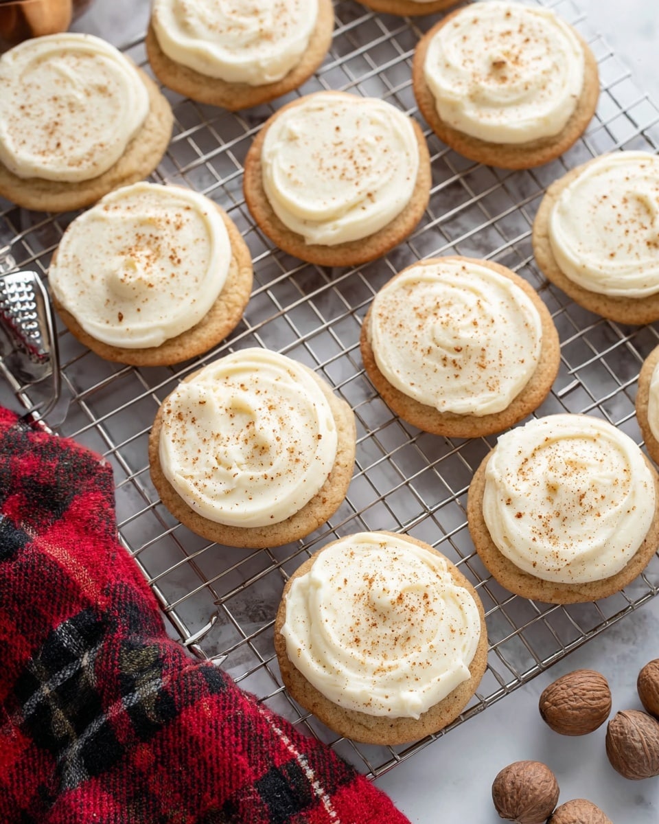 Round cookies with a light golden brown color form the bottom layer. Each cookie has one layer of creamy white frosting spread thickly on top with a soft texture and swirled pattern. The frosting is sprinkled with a light dusting of brown spice that adds a delicate touch. The cookies are arranged on a silver cooling rack over a white marbled surface. Next to the rack, there are whole brown nuts and a silver nut grater. A red and black checkered cloth is partially visible at the bottom left. photo taken with an iphone --ar 4:5 --v 7