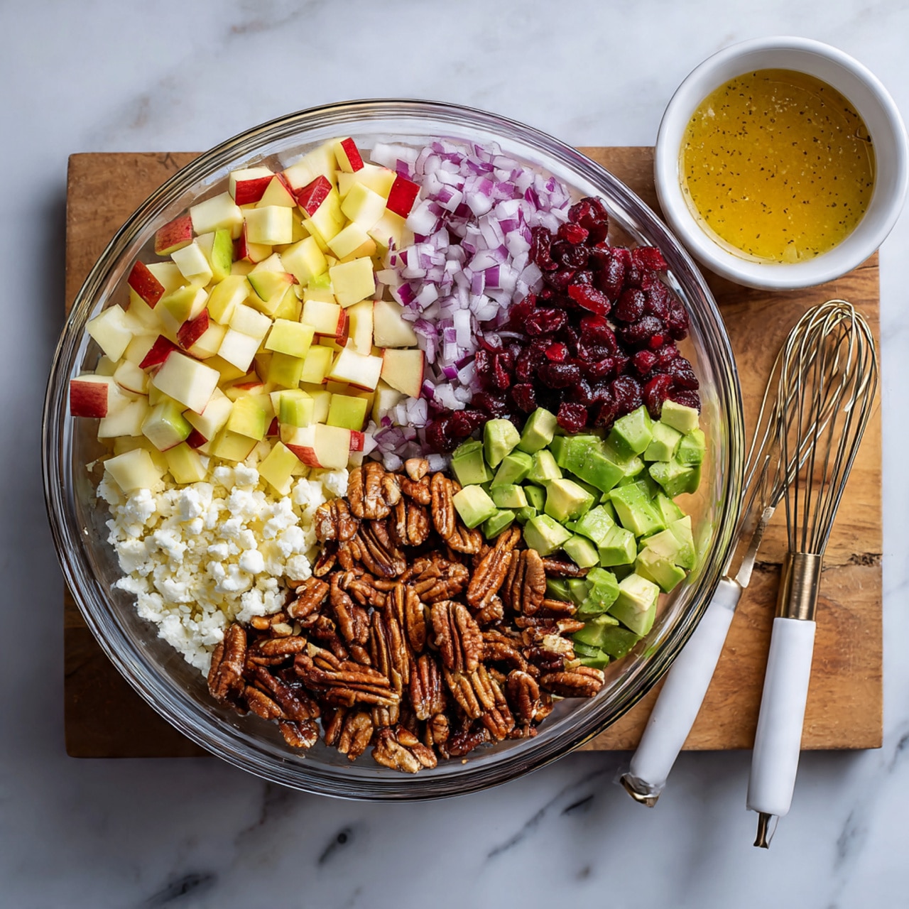The image shows a clear glass bowl with six distinct layers of colorful salad ingredients arranged side by side. Starting from the top left and moving clockwise, there are light yellow and red chunks of apple, dark red dried cranberries, small pieces of purple onion, a white crumbly layer of cheese, light green diced avocado, a cluster of brown pecans, and beige chickpeas completing the circle. Next to the bowl is a small white bowl holding golden brown dressing with a small whisk inside. A set of white and gold serving forks rest on a white marbled textured board beneath the bowl, and the entire scene is set on a white marbled surface. photo taken with an iphone --ar 4:5 --v 7