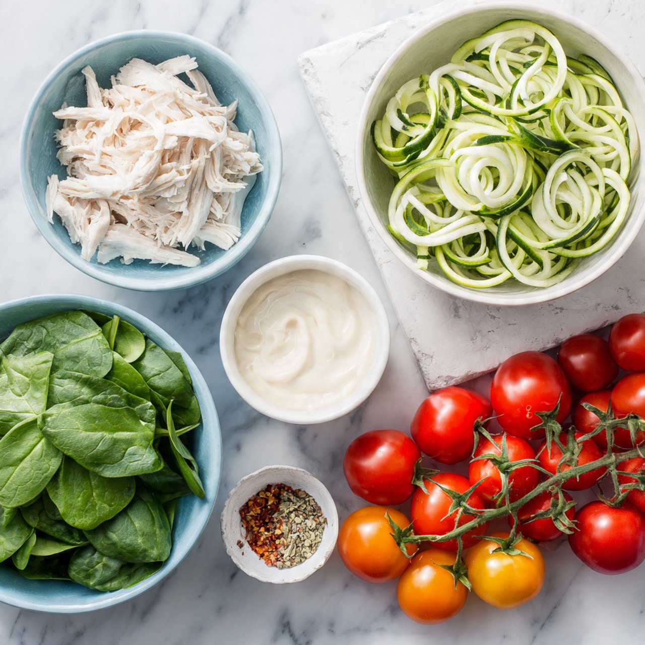 A top view of fresh ingredients laid out on a white marbled texture surface, featuring a white bowl filled with pale green spiralized zucchini noodles in the top right corner. To the left, two light blue bowls hold bright green fresh spinach leaves and white shredded chicken, arranged side by side near the top and center. Below and slightly to the right of the spinach is a small white bowl with creamy white sauce. Near the center is a small bowl with a colorful mix of spices, and across the bottom right of the image is a vibrant cluster of red and orange cherry tomatoes still on their green vine. Photo taken with an iphone --ar 4:5 --v 7