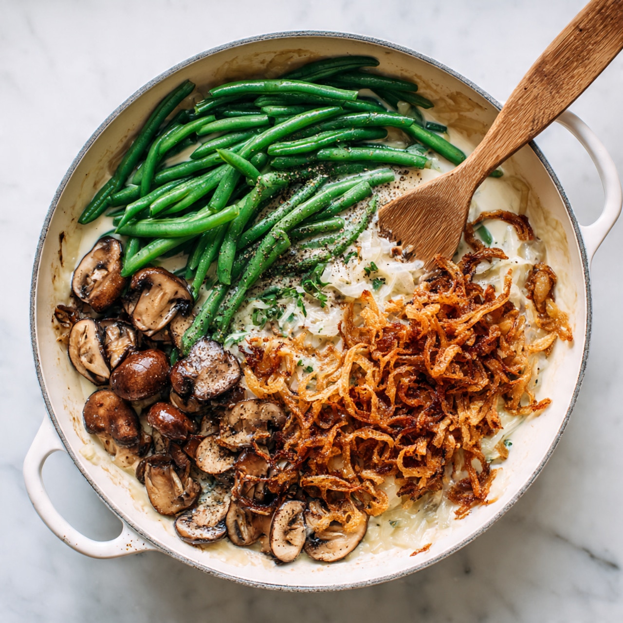 In a white pan on a white marbled surface, there are several layer details: bright green cut green beans cover most of the left and upper side, creamy white sauce with bits of mushrooms fills the lower left area, and sliced brown mushrooms form a small cluster on the lower right. Crispy fried onions in orange-brown color are sprinkled on top near the center, and a light sprinkle of black pepper adds fine dark specks over the dish. A wooden spatula is inside the pan resting over the sauce and vegetables. Photo taken with an iphone --ar 4:5 --v 7