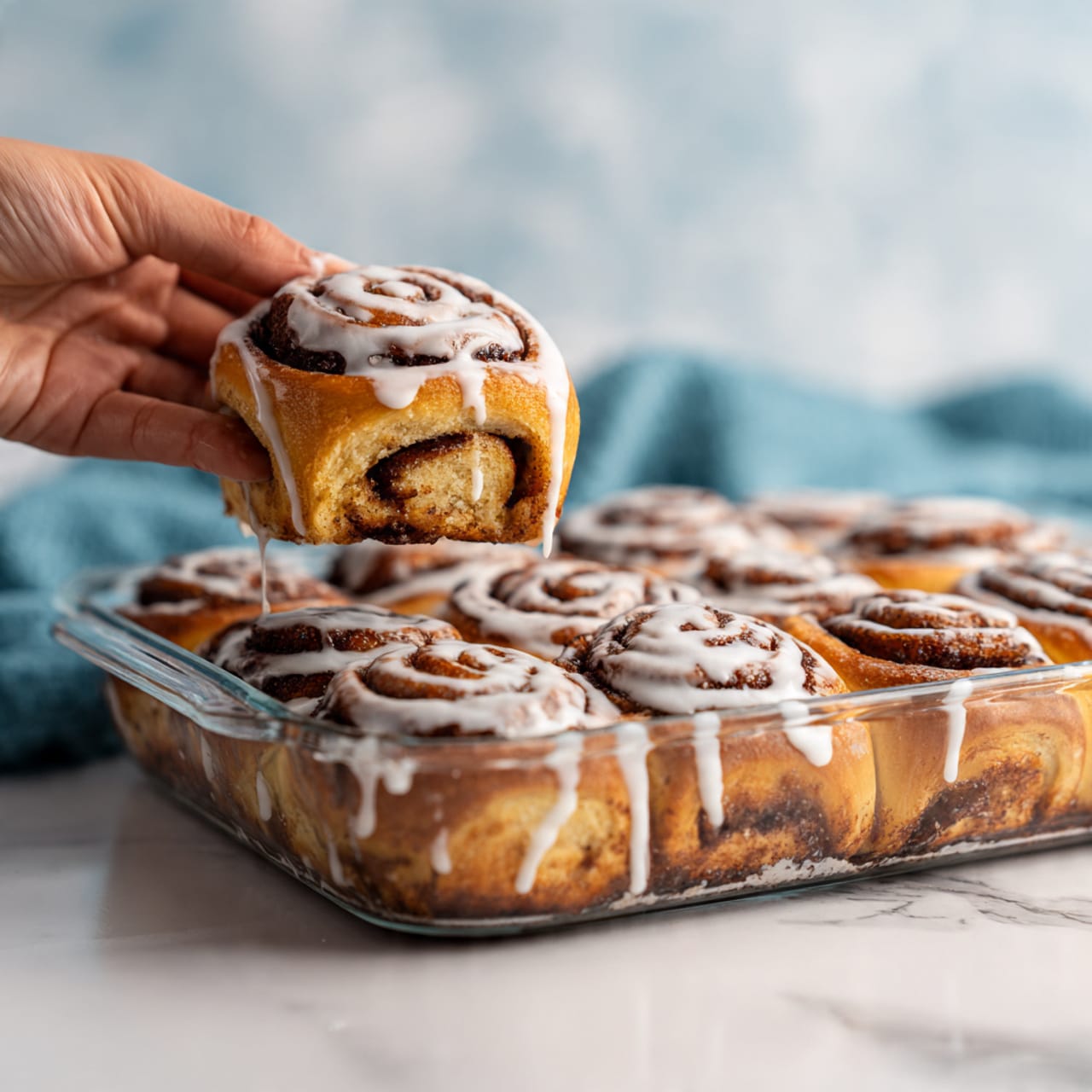 A glass baking dish filled with soft, round cinnamon rolls covered in white icing. Each roll has multiple swirled layers of light golden dough and dark cinnamon filling, topped evenly with creamy white glaze that looks shiny and smooth. A woman's hand is lifting one roll from the dish, showing its thick layers and the shiny icing dripping slightly. The background has a white marbled texture with a blue cloth in the upper left corner. photo taken with an iphone --ar 4:5 --v 7