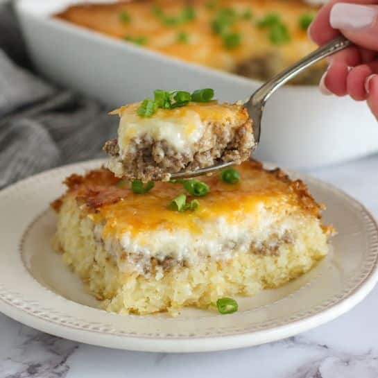 The dish shows a layered casserole on a white plate with a decorative rim, placed on a white marbled surface. The bottom layer is a golden-yellow crumbly base, topped with a crumbly cooked ground meat layer mixed with small bits of orange and white. Above that, there is a creamy white layer with melted cheese that has a golden melted top with small green onion pieces scattered over it. A spoon holds a scoop showing all the layers and green garnish, with a woman's hand holding it. The lighting highlights the textures and colors clearly. photo taken with an iphone --ar 4:5 --v 7