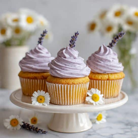 Three cupcakes stand on a white cake stand with a fluted base, each cupcake having a light yellow base with ridged white paper liners. On top of each cupcake is a tall swirl of smooth light purple frosting with a small lavender sprig stuck into it. The cupcakes are arranged in a row, with small white and yellow daisy flowers scattered around the stand. The background is a soft white marbled texture, with blurred white flowers in the distance adding a gentle, fresh feeling. photo taken with an iphone --ar 4:5 --v 7