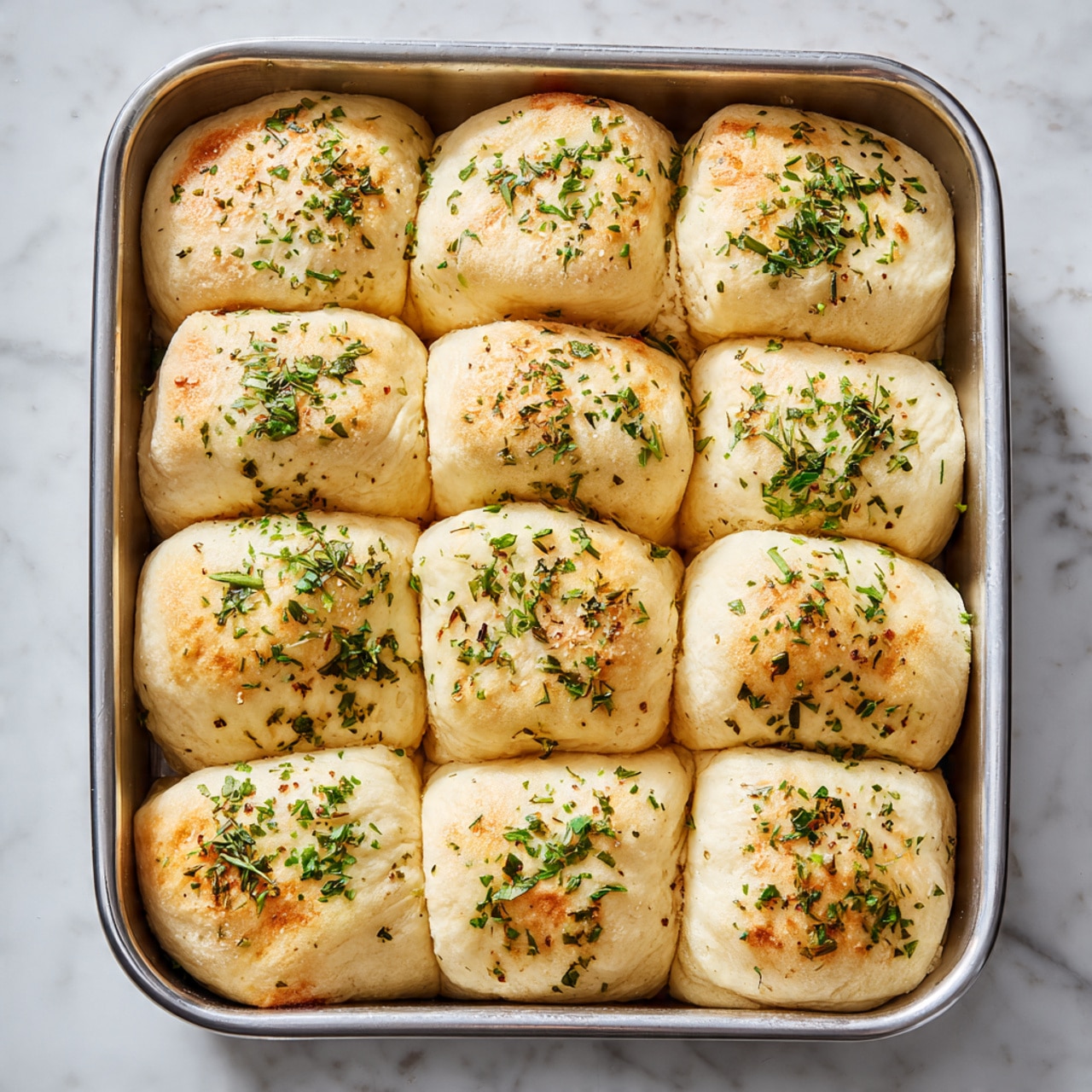 The image shows a metal baking tray filled with 15 dough pieces arranged in three rows of five. Each piece is square with rounded edges, pale beige in color, and topped evenly with small green dried herbs, giving a textured look to the smooth surface. The dough pieces are close together but not touching, showing a soft and fluffy texture ready to rise. The tray sits on a white marbled surface, adding brightness to the image. photo taken with an iphone --ar 4:5 --v 7