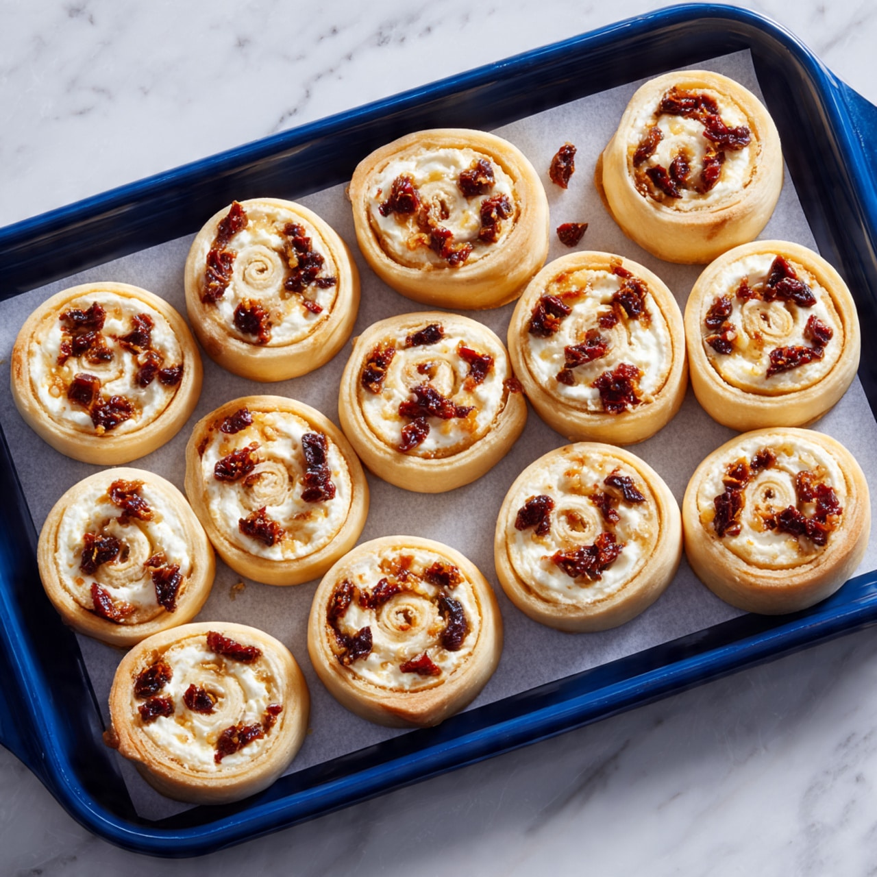 There is a dark blue baking tray with white baking paper on it, holding 21 small spiral pastry rolls. Each roll shows two visible layers: a light golden-brown dough outer layer curled in a spiral, and a creamy white cheese layer with dark brown pieces of sun-dried tomatoes spread evenly inside the spiral. The rolls are placed randomly but close together on the tray, which sits on a white marbled surface. The photo is taken with an iphone --ar 4:5 --v 7