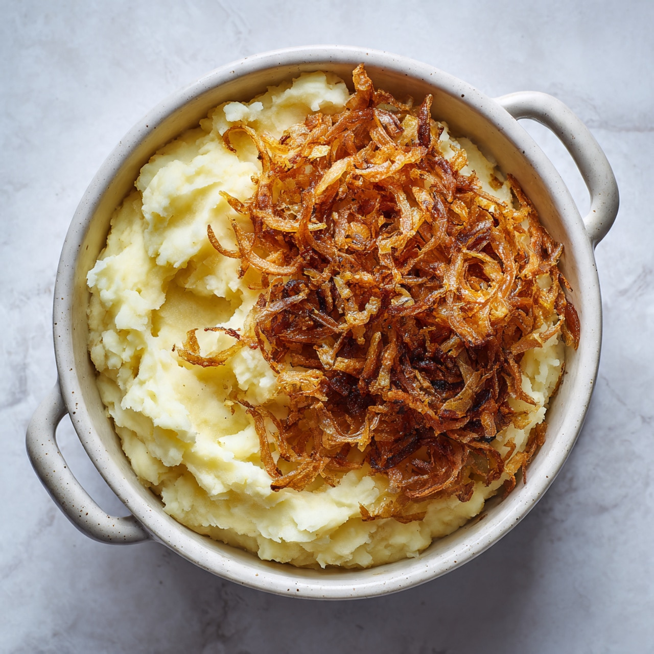 A white pot filled with creamy mashed potatoes forms the base layer, smooth and slightly swirled in texture. On top, there is a generous pile of crispy, golden-brown fried onions mostly gathered on one side, creating a contrast in color and texture. The pot is placed on a white marbled textured surface, and the handles of the pot are visible on either side. The lighting shows a soft natural tone highlighting the textures of the dish. photo taken with an iphone --ar 4:5 --v 7
