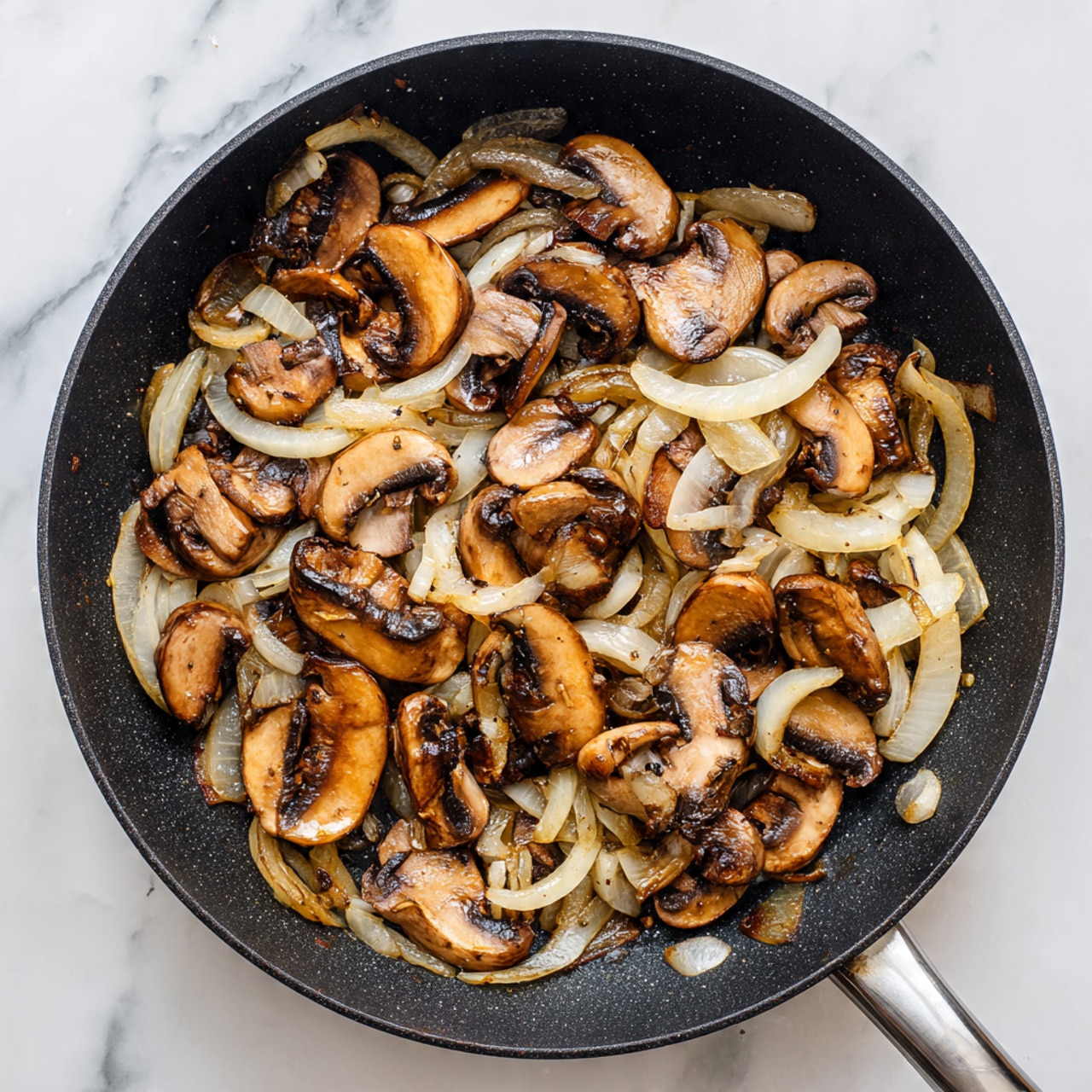 A black cast iron pan filled with cooked mushrooms and onions sits on a white marbled surface. The mushrooms are sliced thickly and show a mix of light to dark brown with some caramelized edges. The onions are cut into thin strips, mostly translucent with light golden-brown spots, spread evenly among the mushrooms. The pan handle extends rightward, and the scene is well-lit with natural light from above. photo taken with an iphone --ar 4:5 --v 7