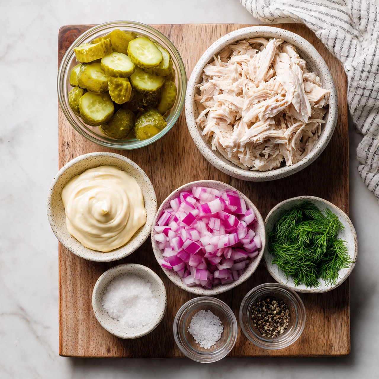 The image shows six small bowls arranged on a wooden cutting board with a white marbled background under it. The largest bowl, placed near the top right, holds shredded white chicken meat with a slightly uneven texture. To its left, a clear glass bowl is filled with diced green pickles showing a glossy and moist texture. Below the chicken, there is a speckled white bowl filled with small diced purple onions, each piece square and firm. To the bottom left of the onions, a small white bowl contains thick, smooth mayonnaise with creamy swirls on top. Above the mayo, a clear glass bowl holds finely chopped fresh dill with a feathery and bright green texture. Lastly, on the lower left side of the board, a small glass bowl contains a mix of salt and pepper with a grainy texture. On the upper right corner of the image, a white and gray striped cloth is partly visible. photo taken with an iphone --ar 4:5 --v 7