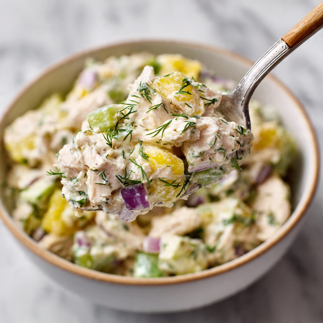 A close-up view shows a silver fork holding a textured scoop of creamy white chicken salad mixed with light green celery pieces, bright yellow pickle slices, small bits of purple onion, and fresh green dill flakes. The background features a blurred view of the same chicken salad in a white bowl with a subtle wooden rim, placed on a white marbled surface. The focus is sharp on the forkful, revealing the creamy and chunky mix with vibrant green and yellow colors standing out against the pale white salad base. photo taken with an iphone --ar 4:5 --v 7