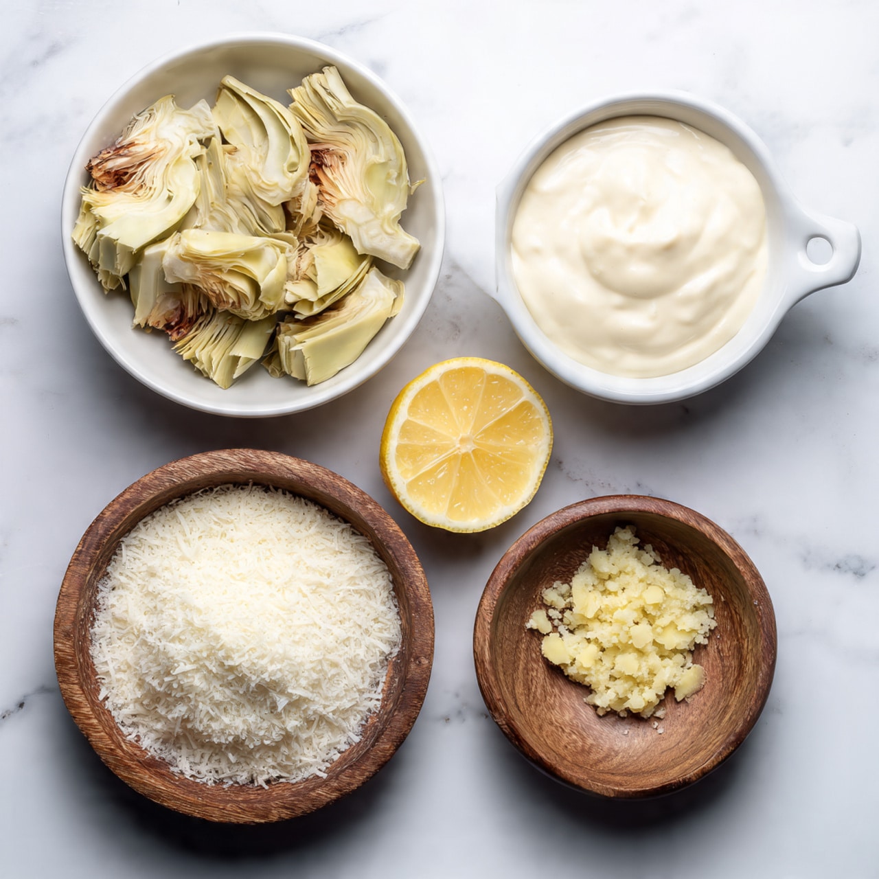 The image shows four bowls and a lemon half placed on a white marbled surface. The top left bowl is white and filled with pale yellow and light brown artichoke pieces. To the right of this bowl is another white bowl with a handle, holding smooth, creamy, off-white mayo-like sauce. Below these two bowls, a wooden bowl filled with finely grated white cheese is on the right. Next to it, a small white bowl contains minced garlic or ginger with a light yellow color. In the center below the artichoke bowl, there is a lemon half with bright yellow flesh facing up. Photo taken with an iphone --ar 4:5 --v 7