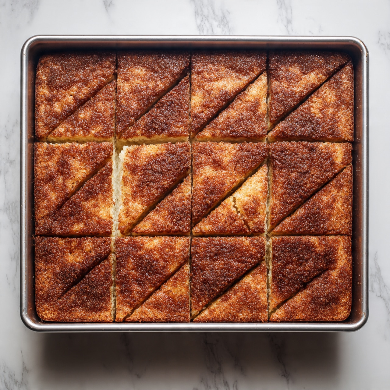 A rectangular metal baking pan filled with a single, thick layer of golden brown baked cornbread topped with a textured layer of cinnamon sugar. The top shows distinct triangle and square shapes where it has been cut, revealing a slightly uneven but soft surface underneath the crunchy cinnamon sugar crust. The pan is set on a white marbled surface, and the image is taken from directly above. photo taken with an iphone --ar 4:5 --v 7