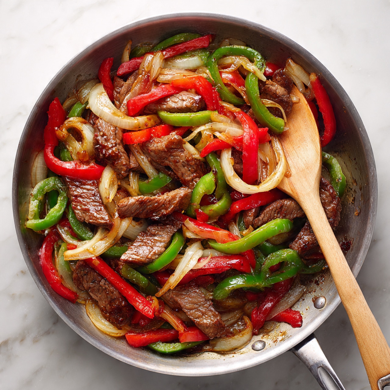 The image shows a close-up of a metal frying pan filled with cooked strips of brown beef mixed with sautéed bell peppers and onions. The bell peppers are red and green, cut into long thin slices, with translucent white onion slices scattered throughout. The beef strips are glossy with a brown sauce coating them, giving a juicy look. A light-colored wooden spatula is stirring the mixture inside the pan. The background is a white marbled texture. Photo taken with an iphone --ar 4:5 --v 7