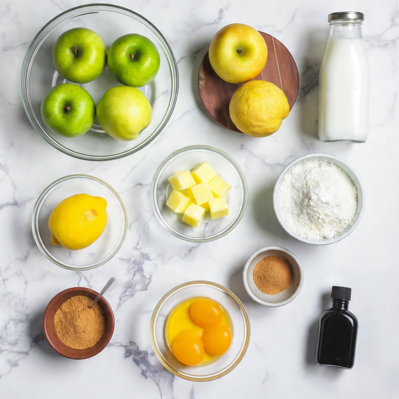 The image shows a flat lay arrangement of ingredients on a white marbled texture, each in clear glass bowls or small brown dishes, except for a white milk bottle in the center. Starting from the top left, two green apples labeled