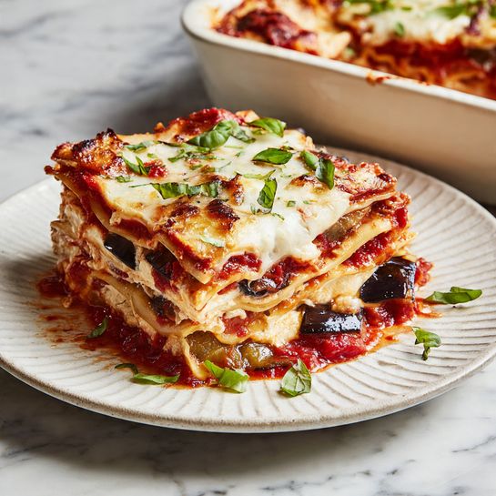 A square piece of lasagna sits on a white plate with a textured edge, placed on a white marbled surface. The lasagna has about five visible layers: a red tomato sauce layer with chunks and bits of dark eggplant, alternating with smooth, light beige pasta sheets layered with melted white cheese on top. Small green basil strips are scattered both on the lasagna and around the plate, adding a fresh touch. The top cheese layer is slightly browned at the edges, creating a soft, melted texture. In the blurred background, a white baking dish holds the rest of the lasagna. Photo taken with an iphone --ar 4:5 --v 7