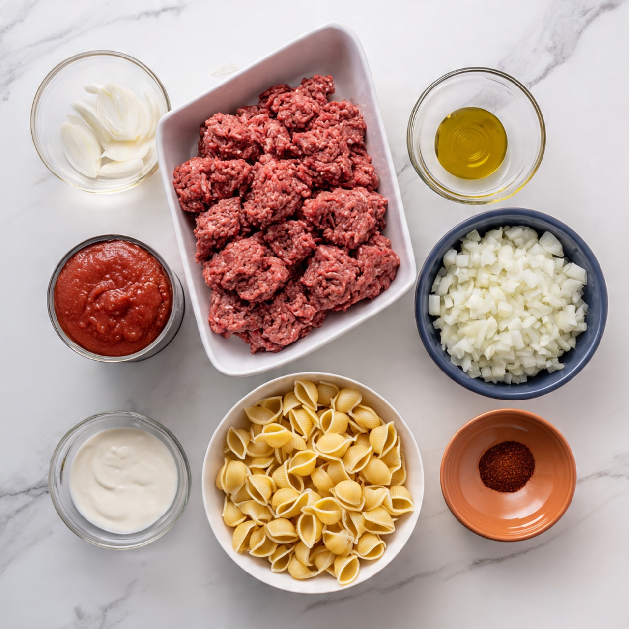 A white tray in the top left holds a large portion of pink ground beef with a label on it. To the right, a white bowl is filled with yellow shell pasta. Below the beef, a small white bowl contains golden olive oil. Next to it, a small dark blue bowl holds red chili powder. To the right of the chili powder, a small orange bowl shows minced garlic with a soft, slightly wet texture. Below is a white bowl filled with roughly chopped white onion pieces. To the left of the onion, an opened can of smooth, red tomato sauce sits with some sauce visible on the can’s edge. At the bottom left and right corners, two clear measuring cups hold white heavy cream and light brown beef broth. All items rest on a white marbled surface. Photo taken with an iphone --ar 4:5 --v 7