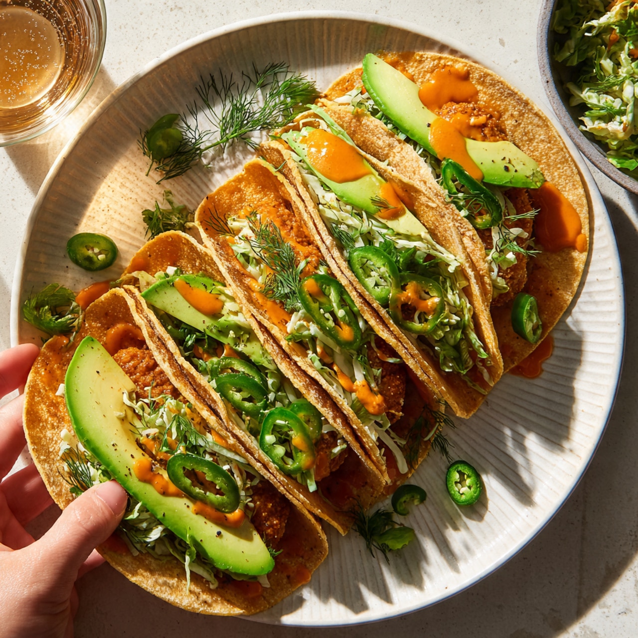 Five round tacos sit on a white plate with a soft edging pattern. Each taco has a golden toasted tortilla as the base layer, topped with a browned, slightly crispy patty. On top of the patties are fresh, thin strips of creamy green cabbage mixed with herbs, placed in loose piles with a light texture. Bright green slices of avocado fan over the cabbage, and fresh green jalapeño slices with visible seeds add a sharp contrast on top. A smooth orange sauce is drizzled over each taco, adding a pop of bright color. Sprigs of fresh dill are scattered across the tacos and on the white marbled surface around the plate. A woman’s hand is holding one taco on the bottom right corner. In the top left corner, there is a partial view of a bowl filled with the cabbage mix, and a glass of a light brown drink is on the bottom left corner. The overall setting is bright with natural light shining down on the tacos showing all details clearly photo taken with an iphone --ar 4:5 --v 7