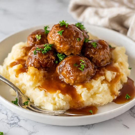 A white plate holds a serving of mashed potatoes topped with five brown meatballs covered in shiny brown gravy. The mashed potatoes form the bottom layer with a soft, creamy texture and light cream color. The meatballs are round and textured, sitting on top of the potatoes. The gravy is thick and glossy, generously poured over the meatballs and some parts of the potatoes. Small green parsley pieces are sprinkled on the meatballs and around the plate for color contrast. Two halves of a meatball on a silver fork rest on the plate near the bottom left. The surface under the plate is a white marbled texture. Photo taken with an iphone --ar 4:5 --v 7