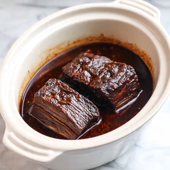 The image shows a white slow cooker filled with a dark, rich stew. Inside the stew are two large, dark brown chunks of meat on the top, with a slightly rough texture and visible fat layers. Beneath the meat, there are several thinner slices of brown pieces, partially covered by the thick, dark brown sauce with a shiny oily surface. The slow cooker has a rounded shape with a small handle visible at the top. The dish sits on a white marbled surface. photo taken with an iphone --ar 4:5 --v 7