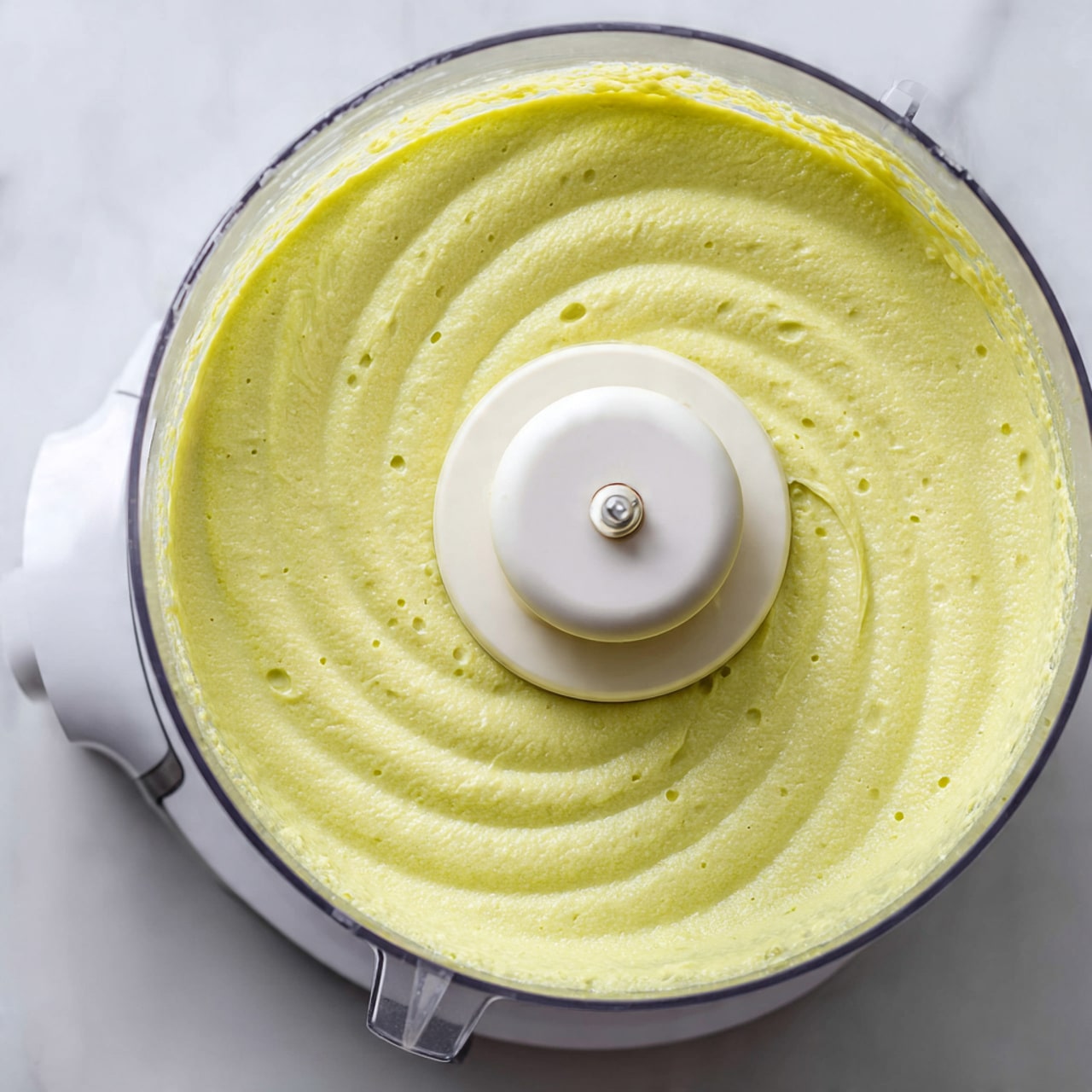 The image shows a close-up top view of a food processor filled with a smooth, creamy light green mixture. The mixture has a slightly thick texture with small bubbles on the surface, evenly spread around the center blade attachment, which is white and round. The clear bowl of the food processor allows the soft green color to be clearly visible. The food processor sits on a white marbled surface. photo taken with an iphone --ar 4:5 --v 7