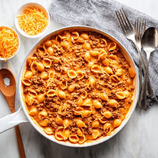 A large white pan mostly filled with orange creamy pasta with small ground meat pieces mixed in, seen on the left side with some empty space on the right showing sauce smeared in swirls; a wooden spoon with dark wood grain rests in the pan over the empty sauce space; near the pan on a white marbled surface are two white bowls filled with the same pasta, one at the top left and one at the bottom right. There are thin shredded orange cheese pieces scattered near the top bowl and two silver forks crossed at the top right corner. A gray and white striped cloth is partially visible under the pan handle. photo taken with an iphone --ar 4:5 --v 7