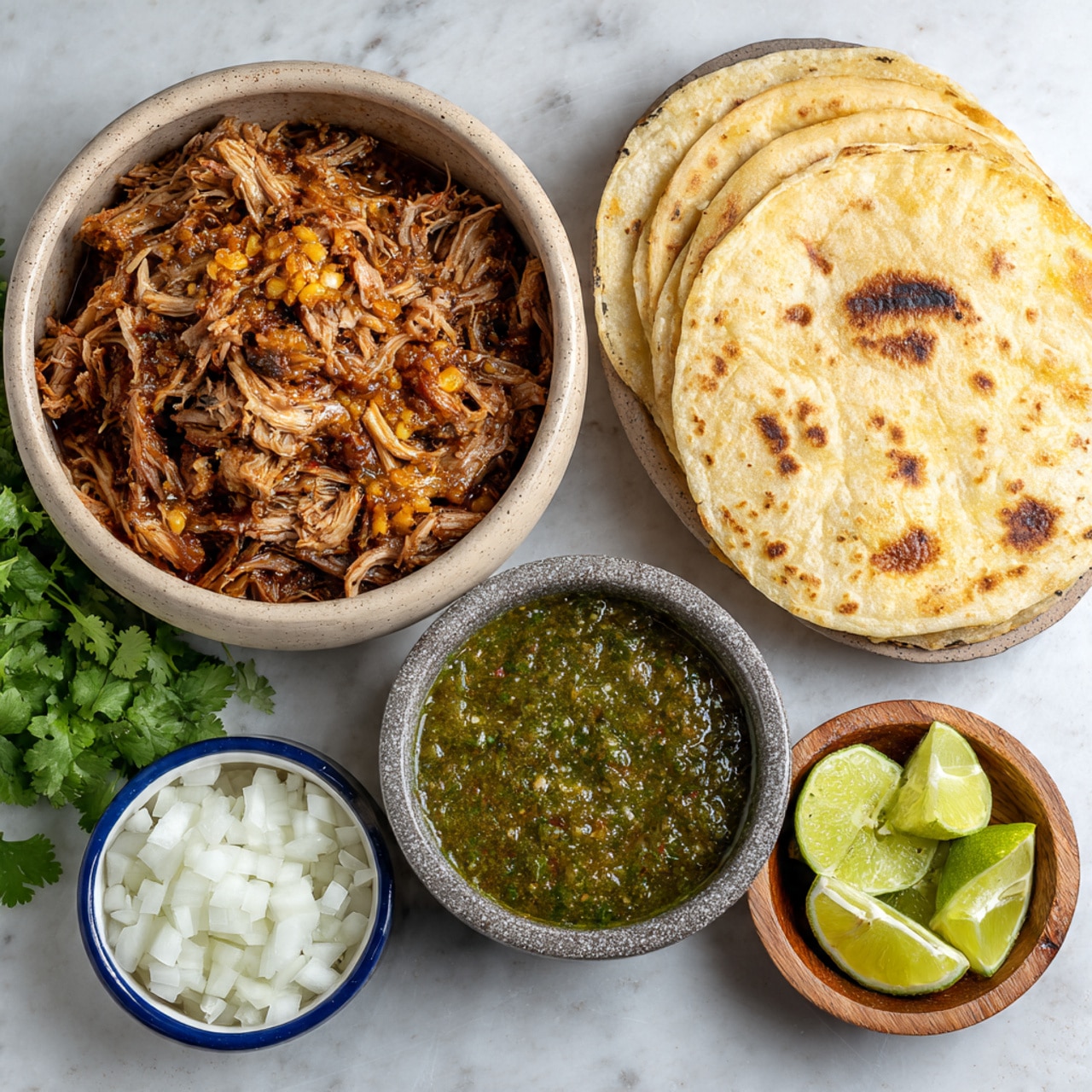 The image shows a meal set on a white marbled surface featuring five separate dishes. The largest is a round beige bowl filled with shredded cooked meat in dark brown and golden tones, placed on the left side near fresh green cilantro leaves. Next to it is a speckled gray bowl full of coarse green sauce with a textured, chunky appearance. On the right, there is a white bowl holding a stack of four toasted tortillas with brown spots and a golden color. Below this is a small white bowl lined with a blue rim, filled with bright green lime wedges. Finally, there is a small wooden bowl containing white diced onions with a smooth texture. photo taken with an iphone --ar 4:5 --v 7