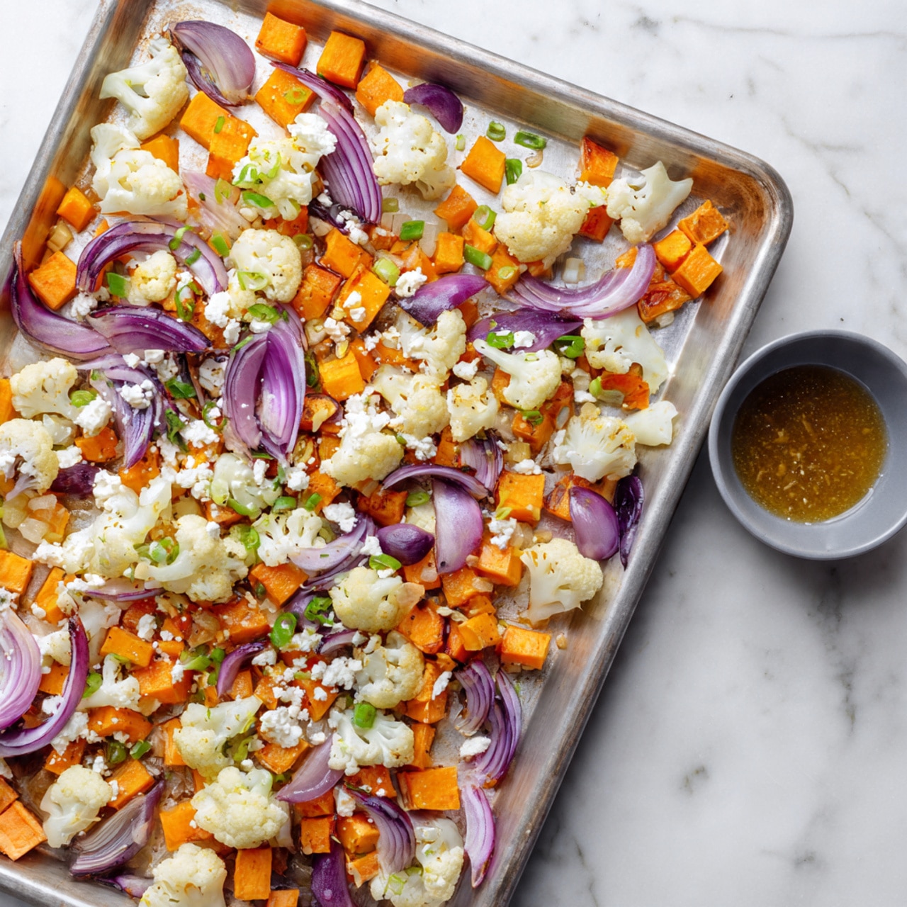 A metal tray filled with several layers of roasted vegetables, mostly orange and white in color. The first layer is small orange cubes that look like sweet potatoes evenly spread across the tray. On top of that, there are roasted cauliflower pieces in white and bright orange arranged all over. Scattered thin slices of purple-red onions add texture and color contrast. Small green pieces of chopped spring onions are sprinkled on top, along with crumbled white cheese. On the tray's upper right corner, there is a gray bowl containing a small amount of amber-colored dressing. The tray is placed on a white marbled surface. Photo taken with an iphone --ar 4:5 --v 7