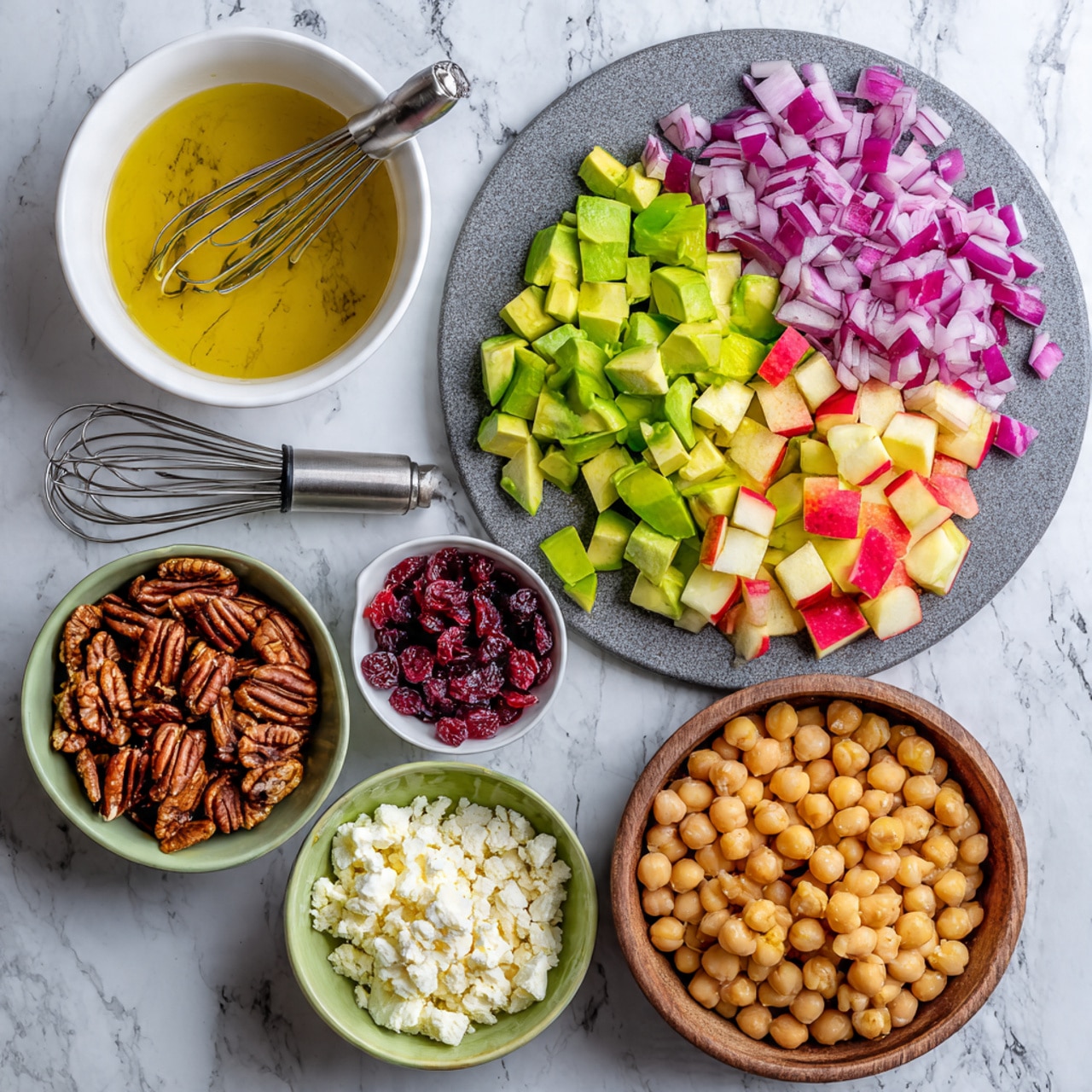 This image shows chopped ingredients for a salad on a white marbled surface. On the right side, three layers of diced food are placed on a round gray cutting board: at the top are small cubes of purple-red onion, below are chunks of bright green avocado, and under that are small pieces of red and yellow apple. To the left of the cutting board, there are five bowls arranged in a loose circle: at the top is a white bowl with a golden-yellow dressing and a small silver whisk inside, below it is a small white bowl with reddish dried cranberries, to the left is a green bowl with brown pecan nuts, below that is a small brown bowl with white crumbled cheese, and at the bottom a large brown bowl filled with light beige chickpeas. Photo taken with an iphone --ar 4:5 --v 7