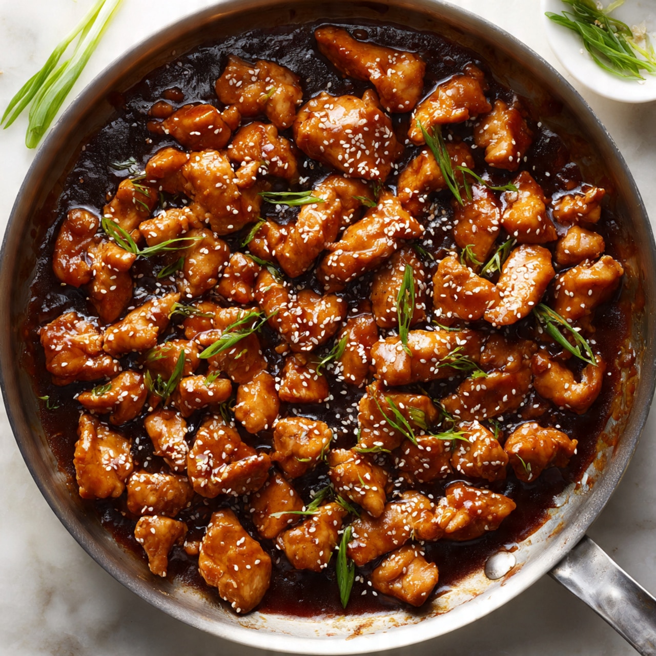 A close-up view of a silver frying pan filled with pieces of glossy, golden brown chicken evenly coated in a thick, dark brown sauce. The chicken pieces are scattered throughout the sauce, each topped with a light sprinkle of white sesame seeds. Small bright green slices of scallions are spread on top, adding a fresh contrast in color. The pan sits on a white marbled surface with some green scallions visible out of focus in the background. The lighting highlights the shiny texture of the sauce and the chicken pieces photo taken with an iphone --ar 4:5 --v 7
