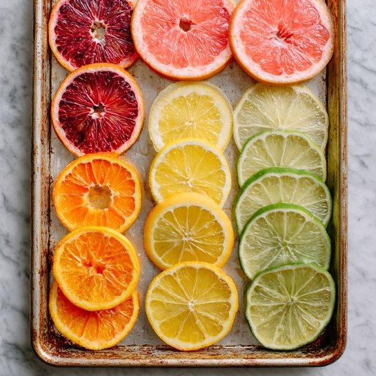The image shows a baking tray with six groups of thinly sliced citrus fruits neatly arranged in a grid. Top left has deep red blood orange slices overlapping in two layers showing textured juicy segments. Top right has two large bright pink grapefruit slices stacked, with visible pulp and thick white rinds. Middle left shows pale yellow lemon slices fanned out in one layer, while middle right has light green lime slices in two loose layers, their translucent flesh and darker green rinds clear. Bottom left has four stacked orange slices with rich orange flesh and bright peels, and bottom right has slightly smaller yellow-orange slices stacked in two layers with a smooth texture. The tray has a worn metallic look and the background is a white marbled texture. Photo taken with an iphone --ar 4:5 --v 7