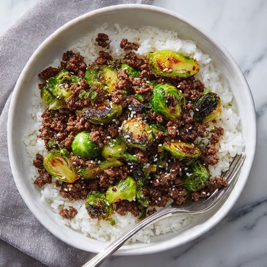 A white bowl filled with a base layer of fluffy white rice, topped with a layer of cooked ground meat mixed with green Brussels sprouts pieces, some with charred leaves showing light green and brown colors, sprinkled with small white sesame seeds. A silver fork rests on the rice at the side inside the bowl. The bowl sits on a white marbled surface with a grey cloth partially visible near the edge. photo taken with an iphone --ar 4:5 --v 7