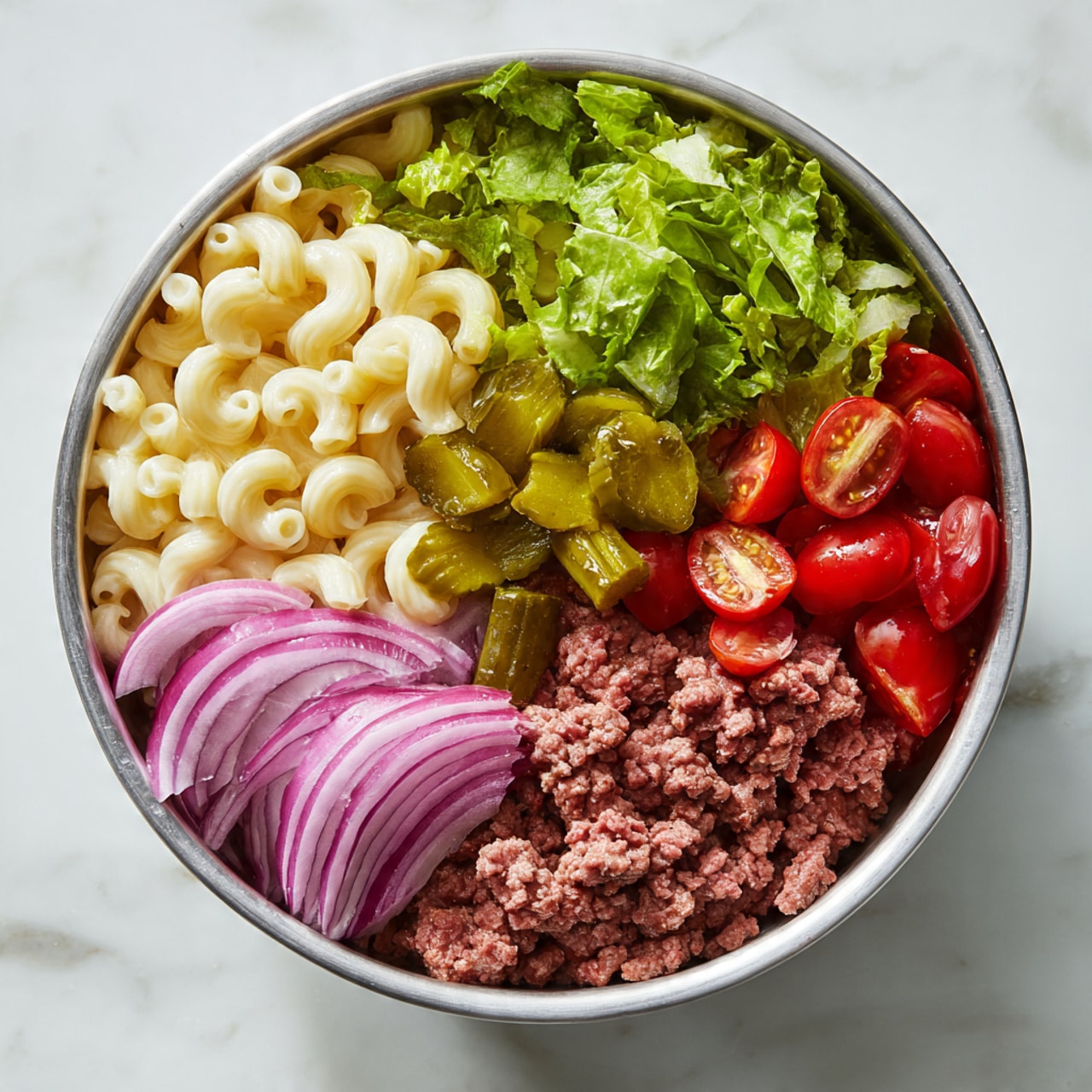 A round silver bowl sits on a white marbled surface filled with layers of colorful ingredients, arranged side by side in sections. On the left, there is pale yellow macaroni pasta, topped partly with small green pickle pieces. Next to the pasta is a pile of bright green leafy lettuce with a curly texture. To the right of the lettuce are halved red cherry tomatoes with a shiny surface. Below the tomatoes is a portion of ground cooked beef with a crumbly texture. Directly below the beef, there are thick slices of purple and white red onion, with crisp edges. The colors and textures of each ingredient stand out clearly in this fresh and vibrant bowl. Photo taken with an iphone --ar 4:5 --v 7