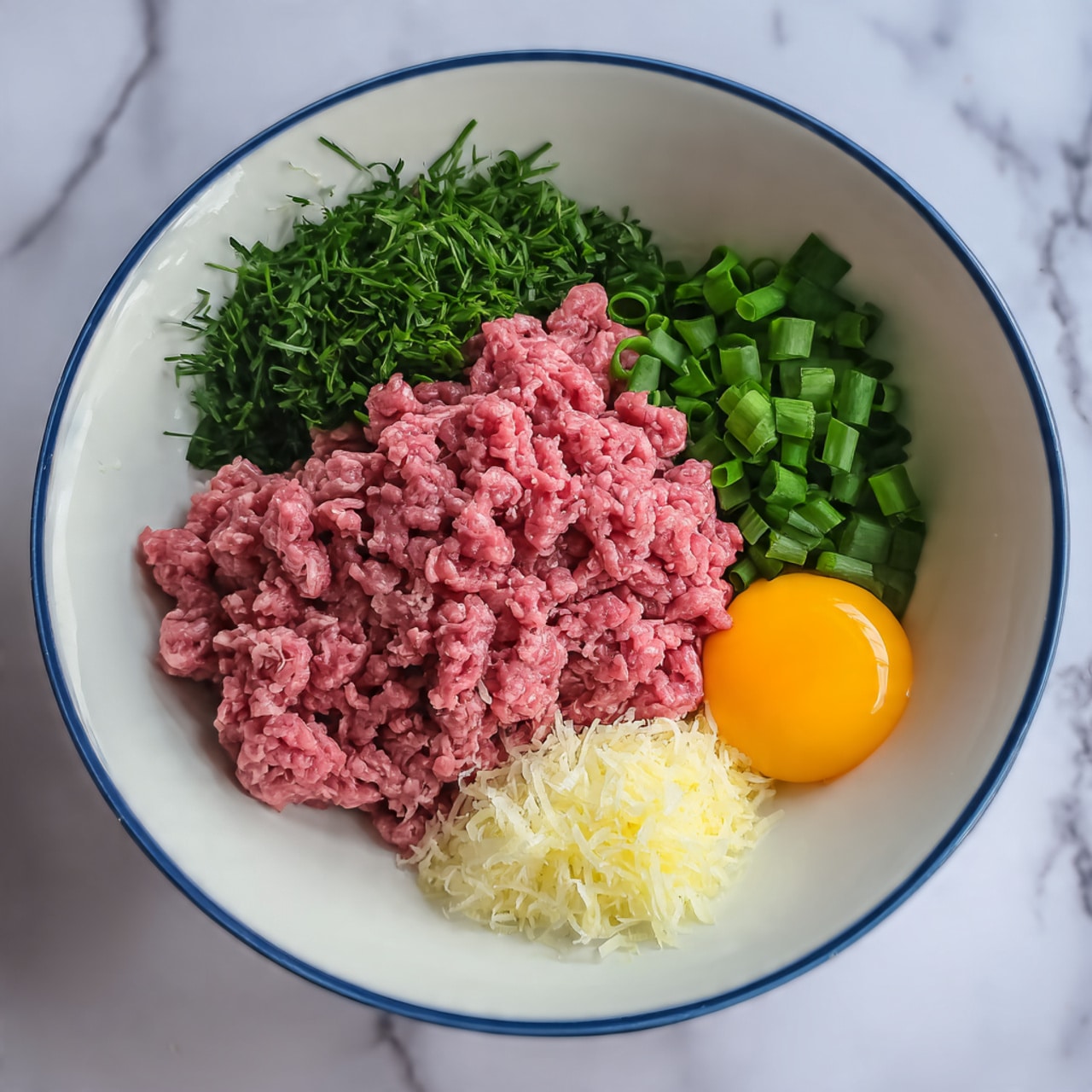 In a large white bowl with a thin blue rim, there are five main layers laid out side by side. The largest layer is finely ground pale pink meat taking up half the bowl on the right. On the top left side is a bunch of fresh green chopped herbs with a fluffy texture. Below that is a pile of sliced light and dark green scallions with a crisp look. Near the center is a small mound of finely grated white garlic or ginger. To the right of the garlic is a dollop of bright orange smooth sauce with a slightly thick and creamy texture. The bowl is placed on a white marbled surface. photo taken with an iphone --ar 4:5 --v 7