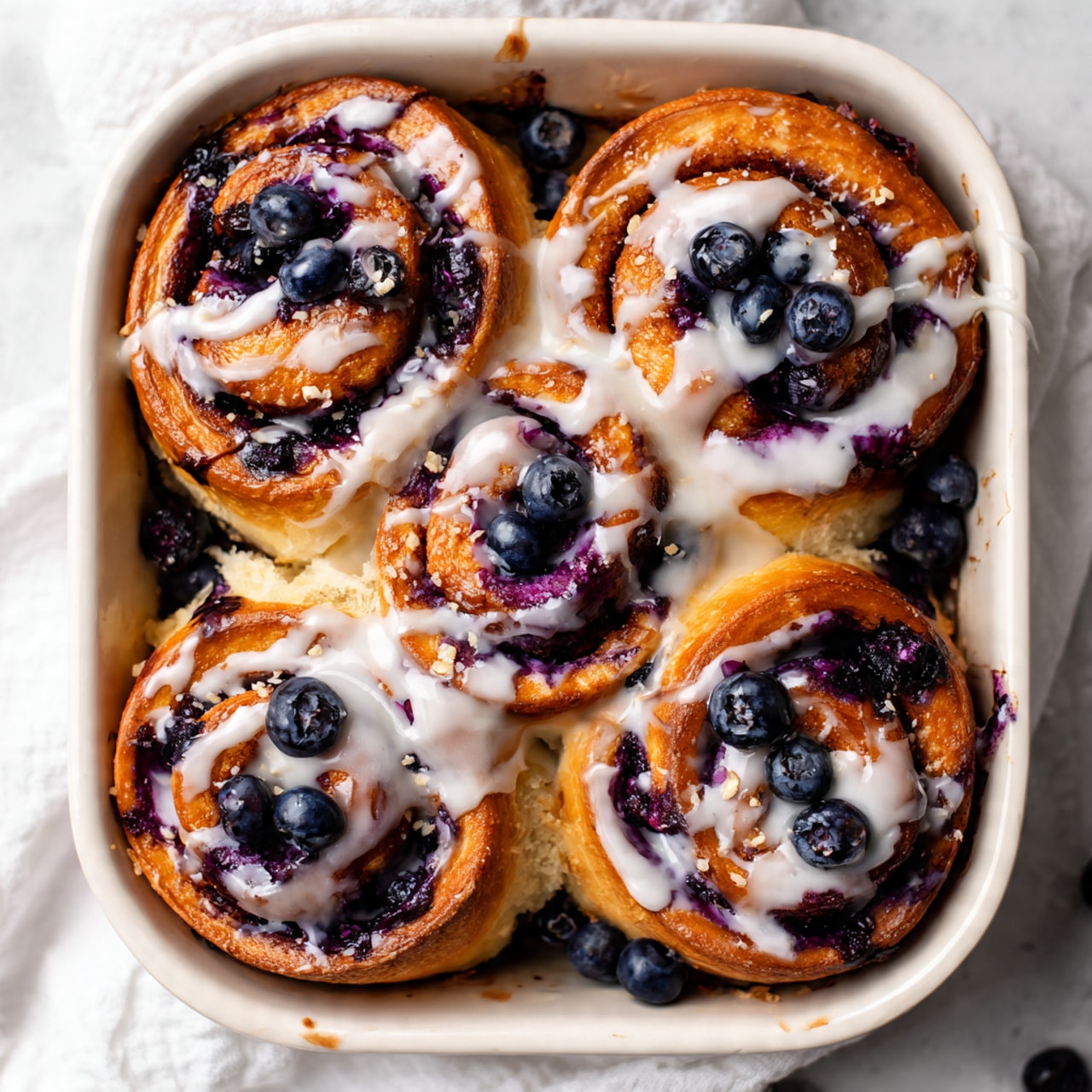 The image shows six golden-brown cinnamon rolls arranged closely in a white baking dish, each topped with a thick layer of white icing that drips slightly down the sides. The rolls have a swirl pattern filled with dark purple blueberry filling, with whole blueberries scattered on top, adding a juicy texture and rich color contrast. The dough appears soft and fluffy, with the blueberry filling oozing out in some areas, giving a glossy sheen. The white marbled surface under the baking dish and a light cloth around the dish add a soft background. Photo taken with an iphone --ar 4:5 --v 7