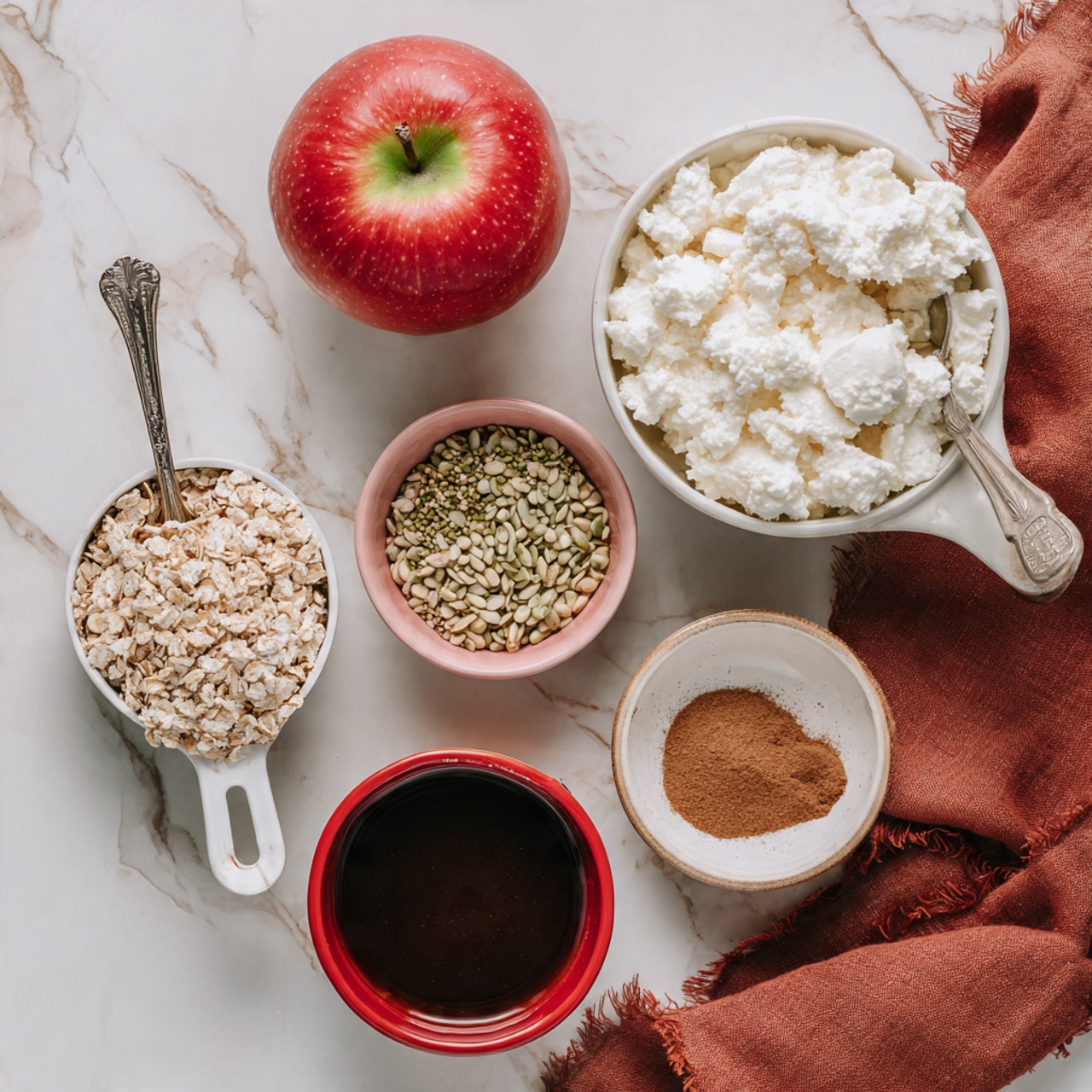 The image shows six main items arranged on a white marbled surface: a red apple with green near the stem at the top left, a small white bowl filled with white cottage cheese and a silver spoon inside on the left below the apple, a white measuring cup filled with light brown granola at the bottom left, a small pink bowl containing a mix of small green and beige seeds at the top right, a small red bowl filled with dark brown syrup or sauce below the pink bowl, and a small white bowl with light brown cinnamon powder near the bottom right. A folded rust-colored cloth is placed at the bottom right corner of the image. Photo taken with an iphone --ar 4:5 --v 7