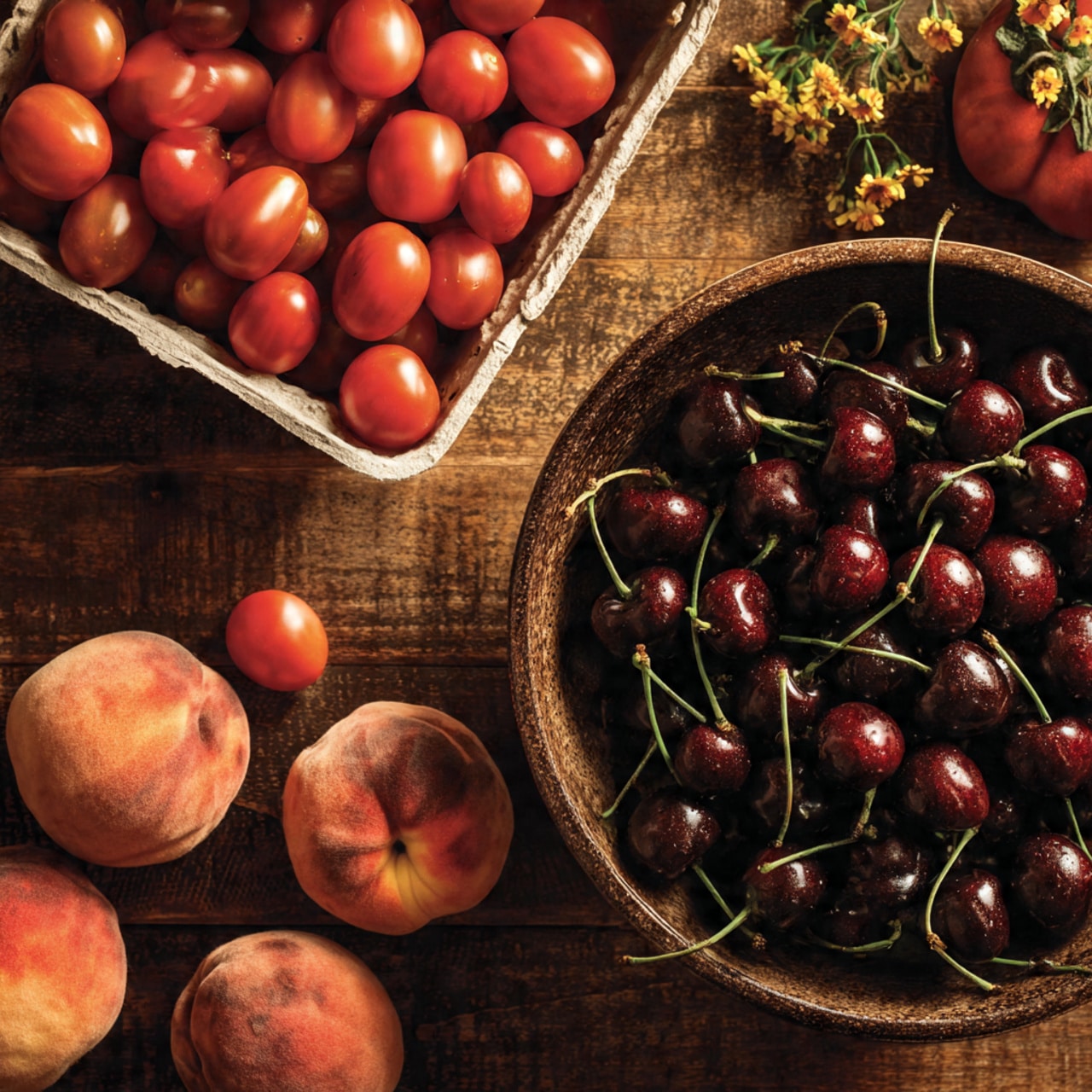 A large round bowl filled with dark red cherries with green stems takes up the right side of the image; the cherries have shiny, smooth surfaces and a few lighter red cherries mixed in. To the left, four peaches with soft fuzzy skin in shades of pink and orange are scattered on a dark wood surface, some with small yellow flowers near them. In the upper left corner, a white rectangular carton holds many bright red cherry tomatoes, some spilling out onto the wood, accompanied by a few tiny yellow flowers. The image shows a warm, natural light with a cozy, rustic feel. Photo taken with an iphone --ar 4:5 --v 7