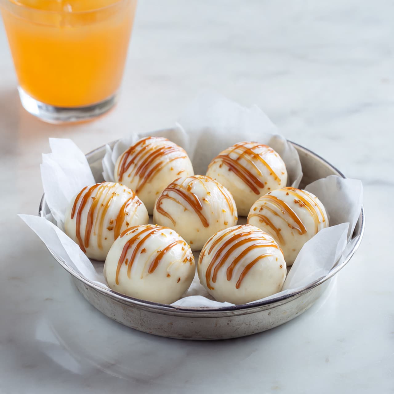 There are seven round balls covered with a smooth, creamy white coating, each ball decorated with four to five thin, random lines of light brown caramel sauce drizzled on top. They are placed closely together in a small silver tart pan lined with white parchment paper. The background is a white marbled surface, and part of a clear glass with an orange drink is visible at the top left corner. Photo taken with an iphone --ar 4:5 --v 7