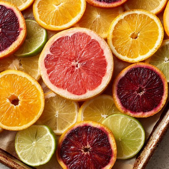 The image shows a baking tray lined with brown parchment paper, holding one large pink grapefruit slice in the center, surrounded by oranges with bright orange segments, deep red blood oranges with dark reddish-purple centers, light yellow lemon slices, and pale green lime slices. The citrus slices are spread out evenly, revealing both the colorful flesh and the thin rinds, creating a mix of vibrant orange, red, yellow, and green shades against the rustic tray backdrop. Photo taken with an iphone --ar 4:5 --v 7
