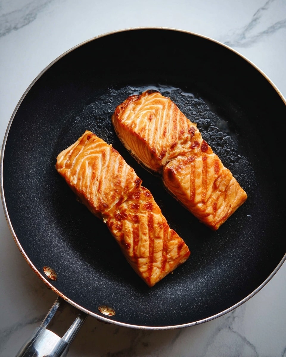 Two pieces of cooked salmon with a golden brown sear and clear grill lines lie side by side on a black frying pan. The salmon's surface shows layers of light orange and pinkish tones with slightly crisp edges. The pan has a silver handle and is set against a white marbled texture background. photo taken with an iphone --ar 4:5 --v 7