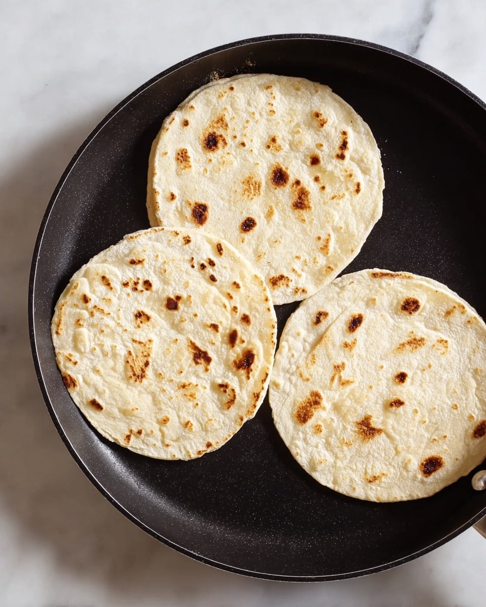 The image shows three round, lightly toasted tortillas on a black pan. Each tortilla has a pale cream color with small toasted brown spots spread unevenly across their surfaces. The tortillas are flat with a slightly rough texture, and they are evenly spaced on the pan. The pan itself is placed on a white marbled surface, which has a smooth and shiny texture. The light in the image highlights the different shades of cream and brown on the tortillas, making them look warm and freshly cooked. photo taken with an iphone --ar 4:5 --v 7