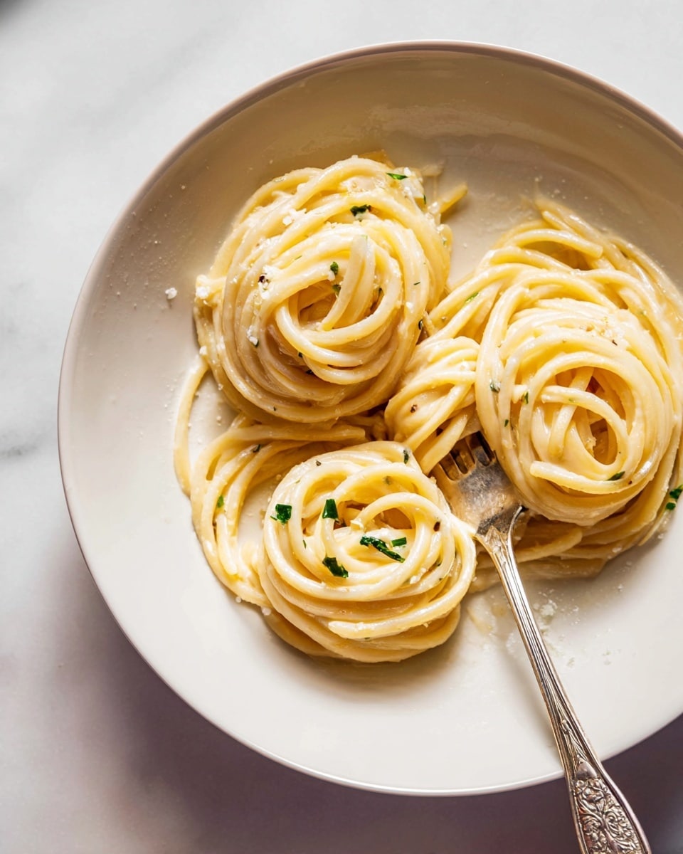 The dish shows thick spaghetti noodles arranged in three small twirls on a white round plate with a smooth surface. The noodles are creamy beige with a light shiny texture, coated evenly with a pale yellow sauce. Small green herb pieces sprinkled lightly over the pasta add a touch of color. A silver fork with ornate details rests inside the plate, partly under the noodles, with its tines pointing toward the center. The plate sits on a white marbled surface, with soft natural lighting highlighting the creamy texture of the pasta. Photo taken with an iphone --ar 4:5 --v 7